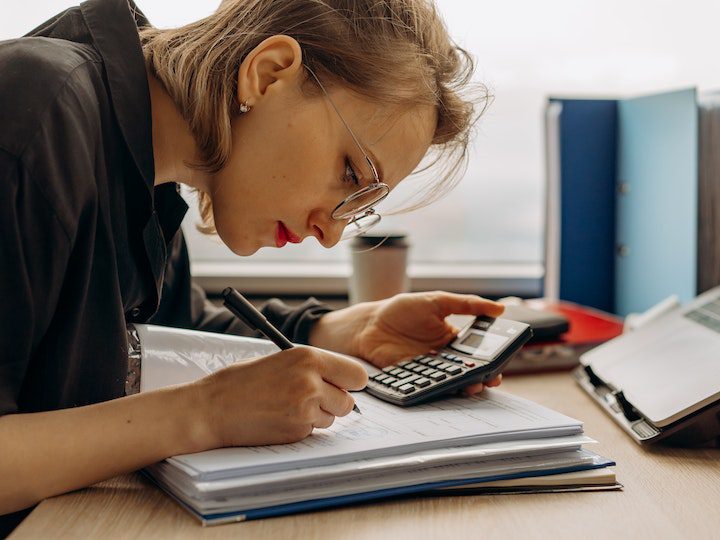 a-woman-writing-on-a-book-while-holding-a-calculator
