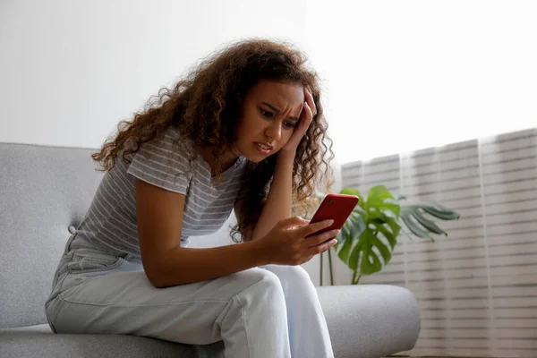 Portrait of young beautiful black woman with depressed facial expression sitting on grey textile couch holding her phone. Cyber bullying victim concept. Sad female in her room. Background, 