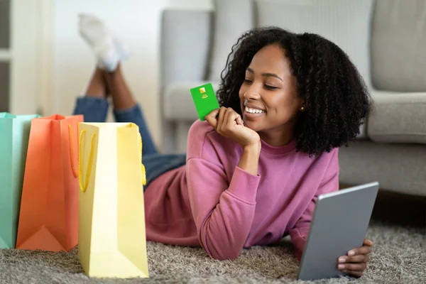 Young black lady holding credit card, shopping online via laptop, lying on floor with gift bags at home. Happy customer purchasing goods in web store, using touch pad