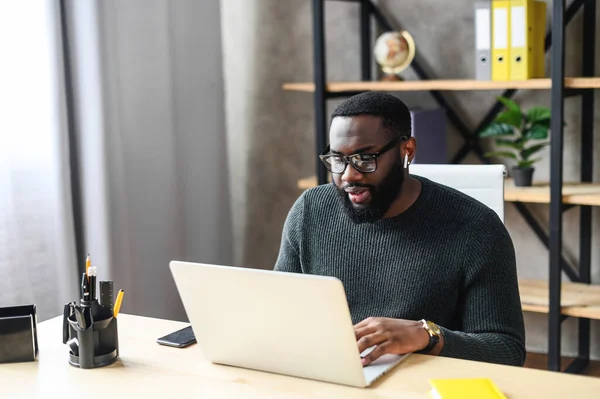 African-American young guy works with laptop while sitting at the office desk, he is in eyeglasses typing on keyboard