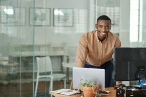 Portrait of a young African businessman smiling while leaning against his desk in an office and working on a tablet