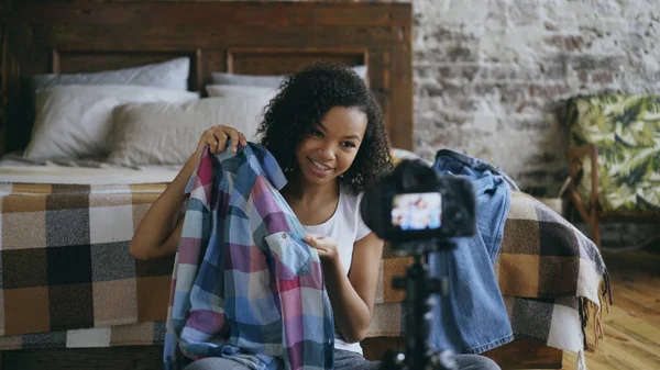 Cheerful mixed race woman sitting near bed recording video blog about her wardrobe for travel with dslr camera at home 