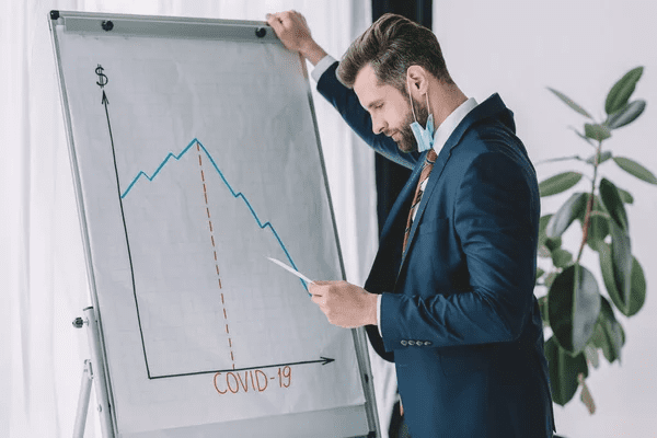 Thoughtful businessman looking at paper while standing near flipchart with covid-19 inscription and graphs showing decrease